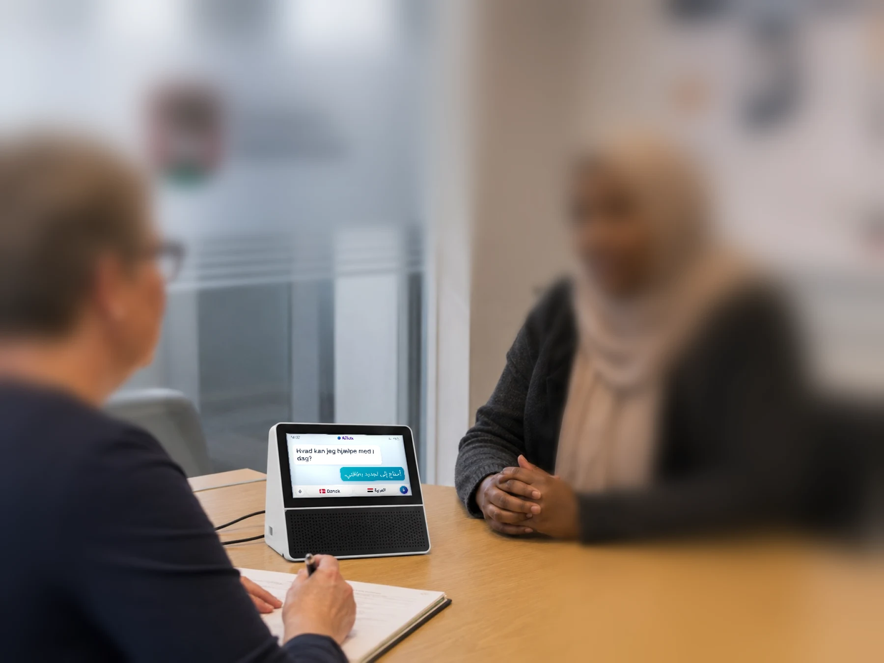 AiTolk-enheden står på bordet i et kommunalt mødelokale mellem en sagsbehandler og en borger. På skærmen står samtalen på dansk og arabisk.